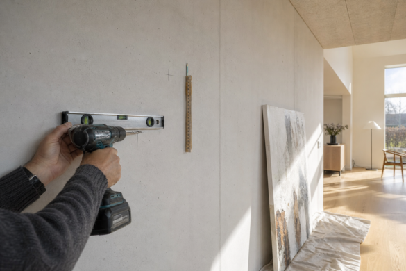 Hænder med borehammer og vaterpas ved en stor hvid væg i et nybygget hus, mens et stort maleri står lænet op ad væggen og venter på ophæng